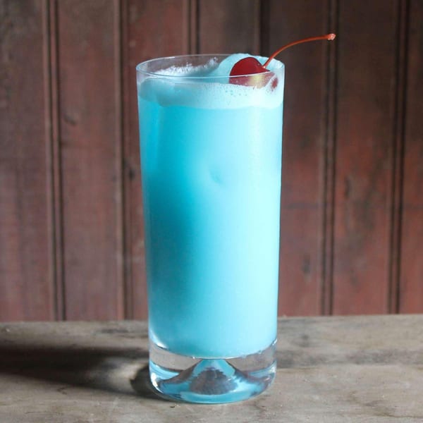Tall glass of vibrant blue cocktail with foam and a red cherry garnish against a wooden background.