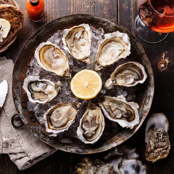 A platter of fresh oysters on ice with a lemon slice, hot sauce, and a drink.