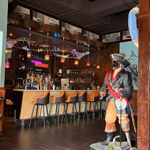 A pirate statue stands beside a bar with orange stools, neon lights, and a TV screen.