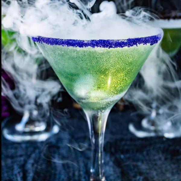 A vibrant green cocktail with a blue sugared rim, emitting dramatic white smoke from the glass.