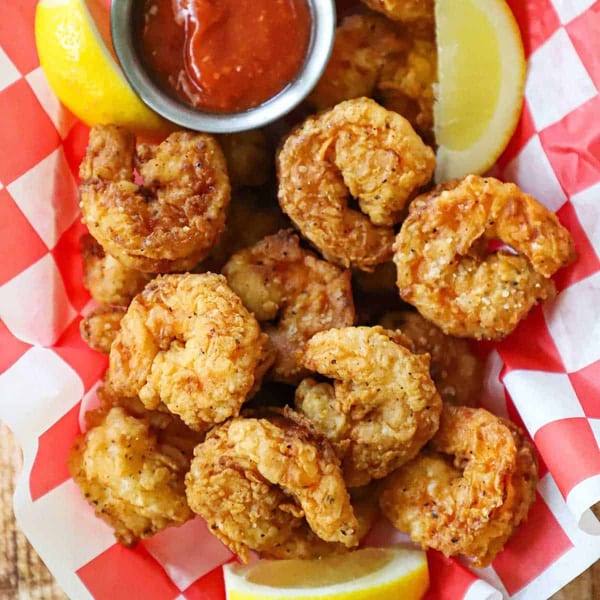 Crispy fried shrimp with lemon wedges and a side of red dipping sauce in a checkered basket.
